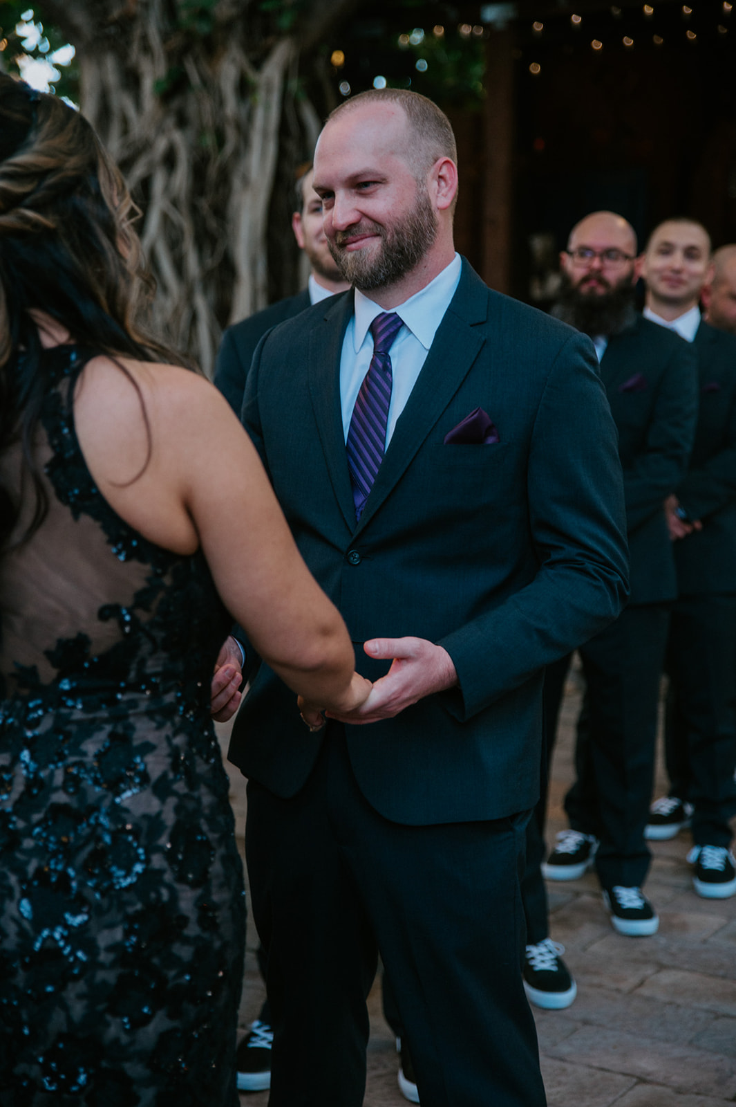 Groom smiling as he holds his partner's hands during the ceremony while his groomsmen look on.
