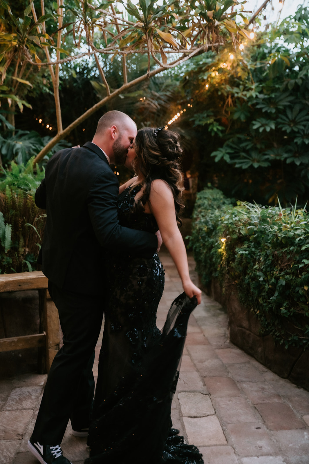 Couple sharing a romantic kiss along a lush greenhouse pathway at boojum tree hidden gardens.
