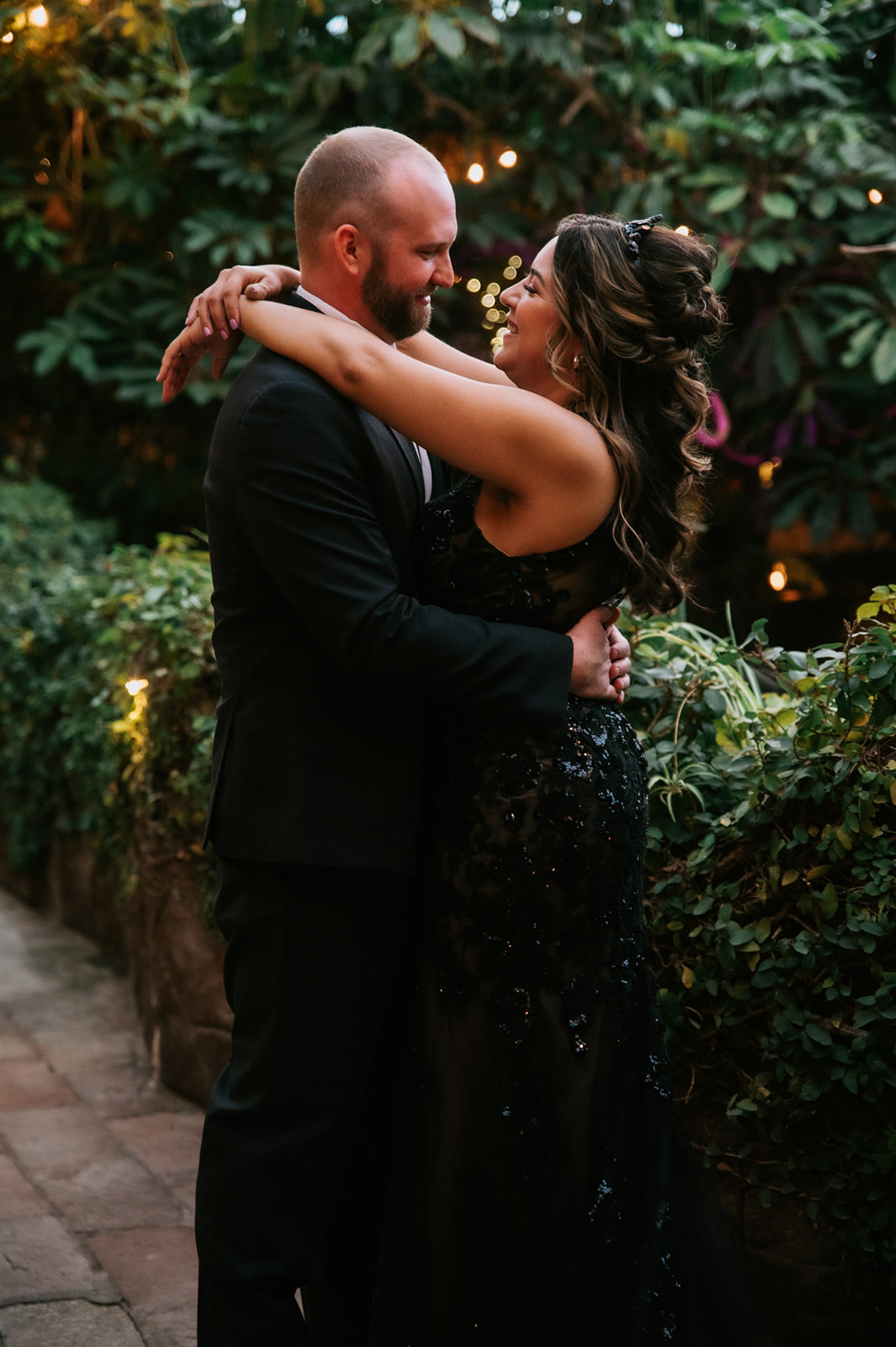 Bride and groom embracing beneath glowing garden lights during romantic wedding portraits.
