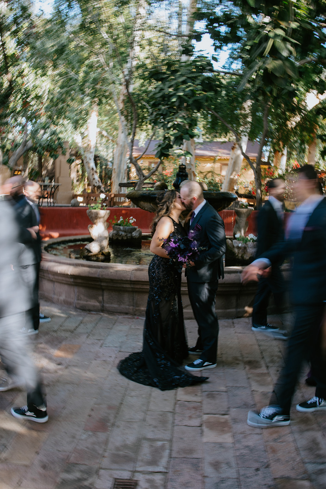 Couple sharing a kiss beside the courtyard fountain with guests captured in creative motion blur at boojum tree hidden gardens.
