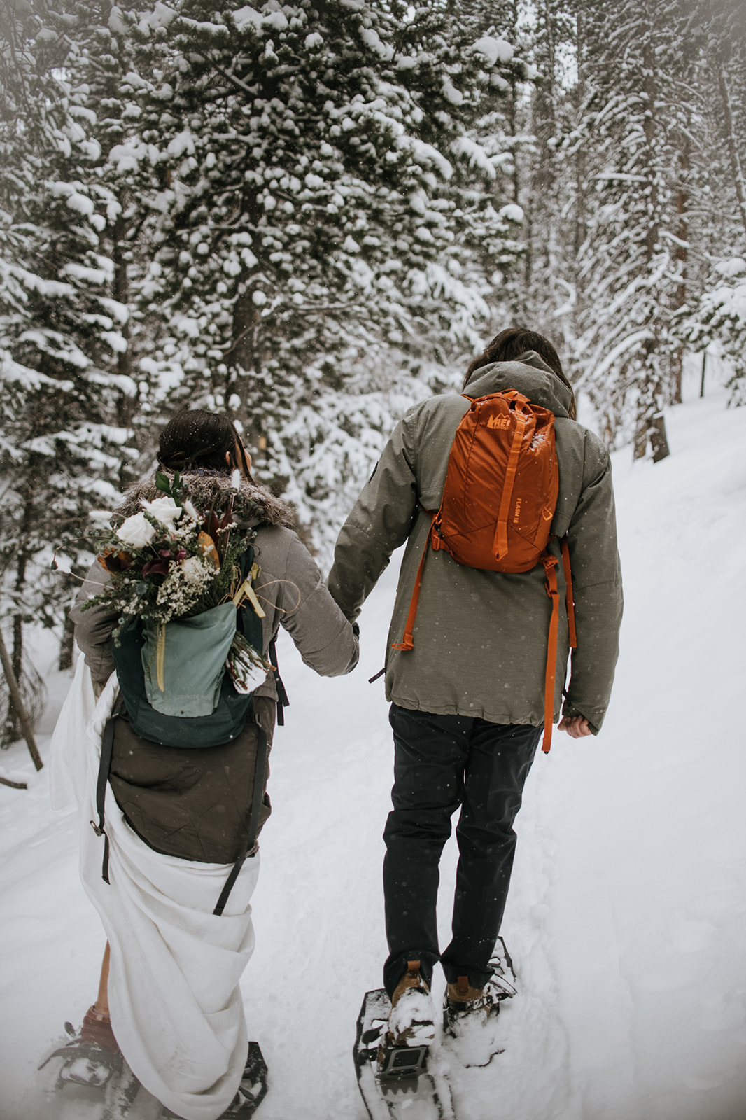 Couple hiking hand in hand through snowy trees during a winter rocky mountain national park elopement