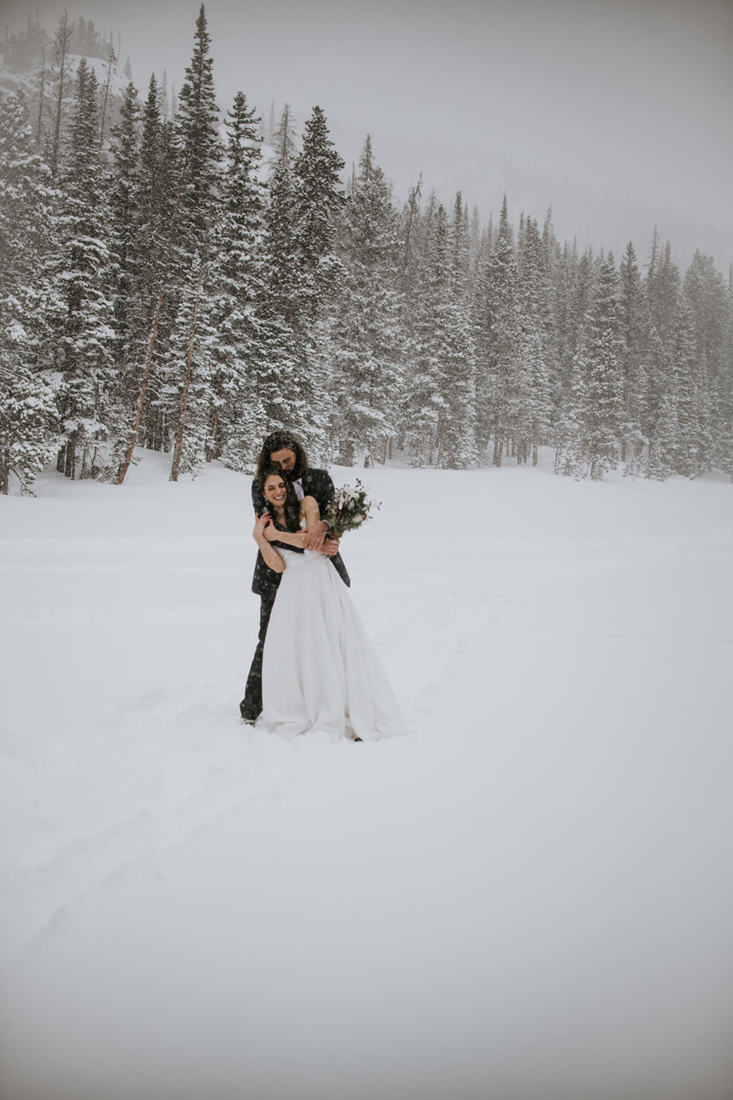 Wide shot of couple standing together on a frozen lake during a winter elopement surrounded by pine trees