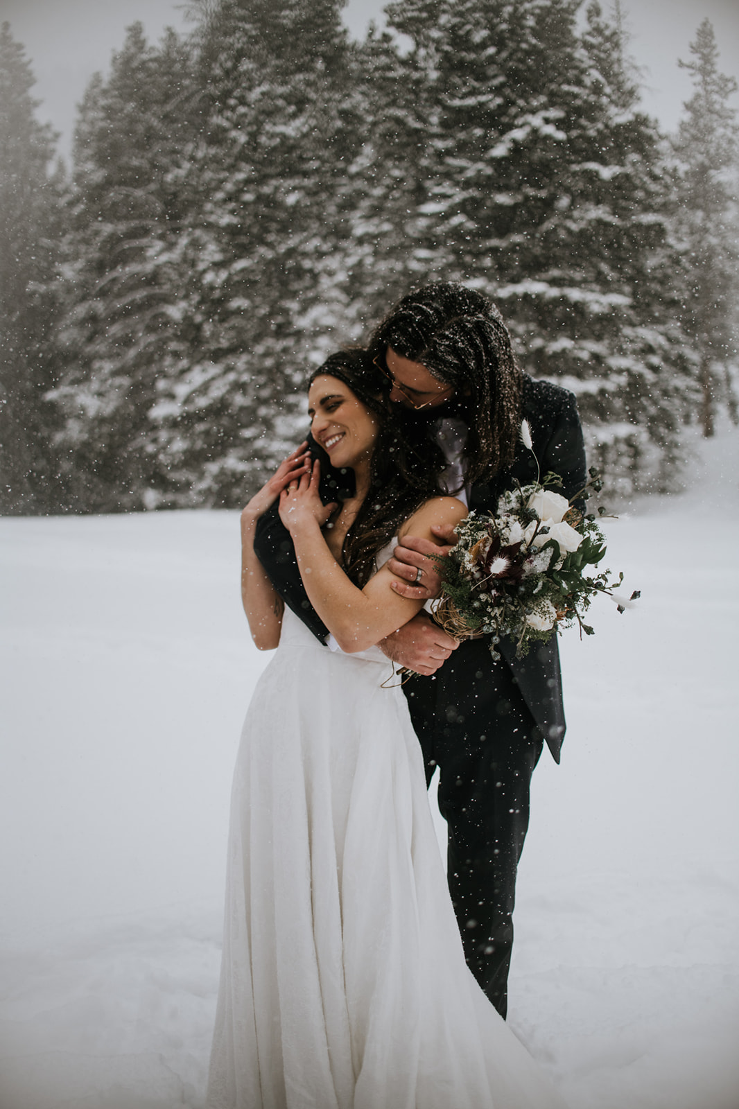 Bride smiling as groom hugs her from behind during a snowy mountain elopement