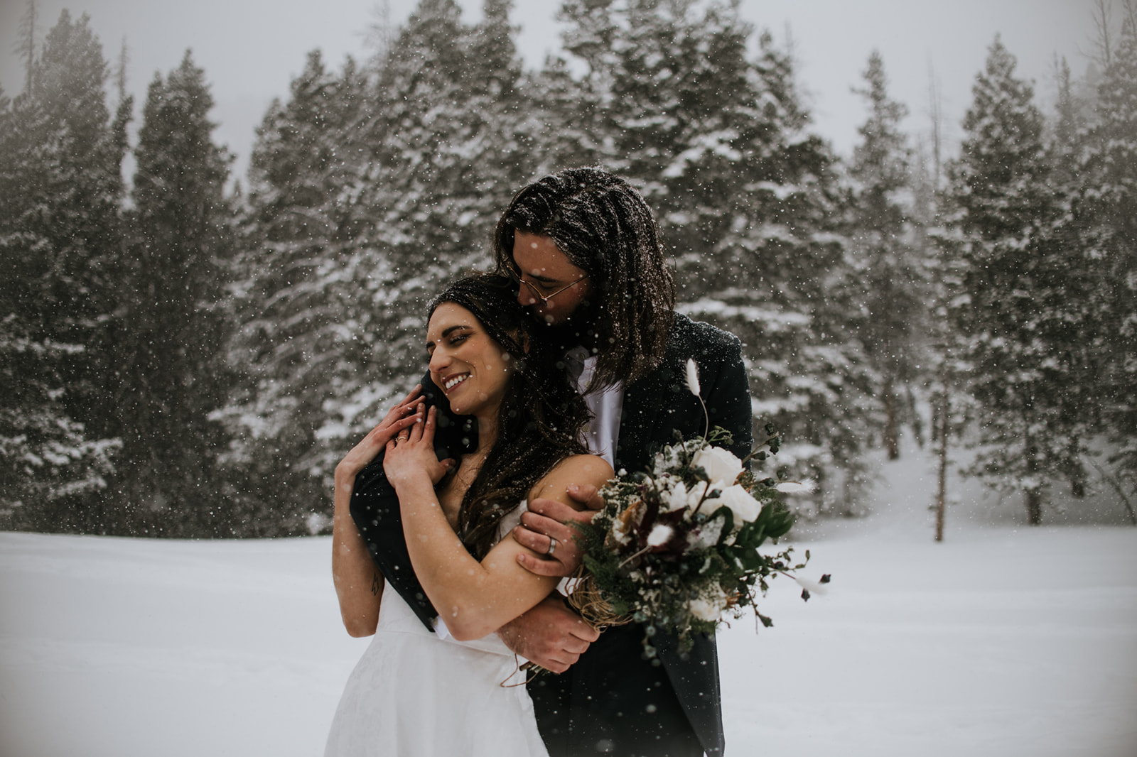 Close-up of bride smiling as groom hugs her from behind during a snowy mountain elopement