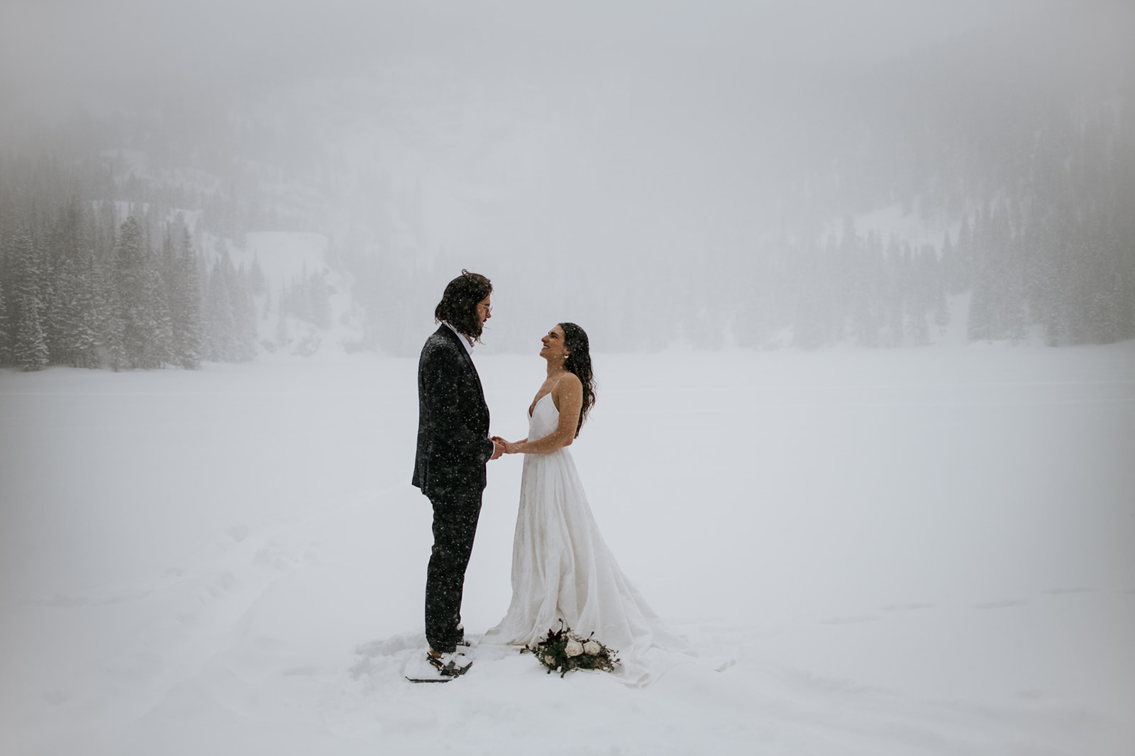 Bride and groom holding hands in falling snow during a rocky mountain national park elopement on a frozen lake