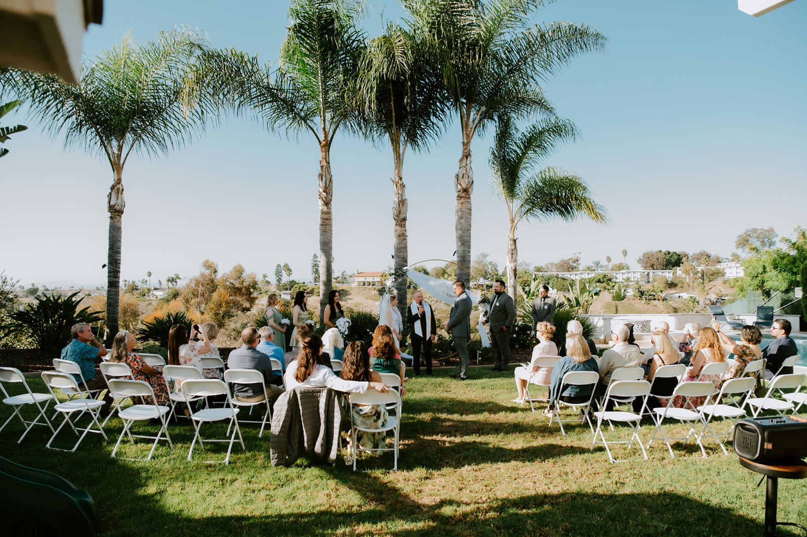 Outdoor ceremony with guests seated on white chairs beneath palm trees, a backyard-style setup that shows how to plan an elopement with family in a relaxed setting.