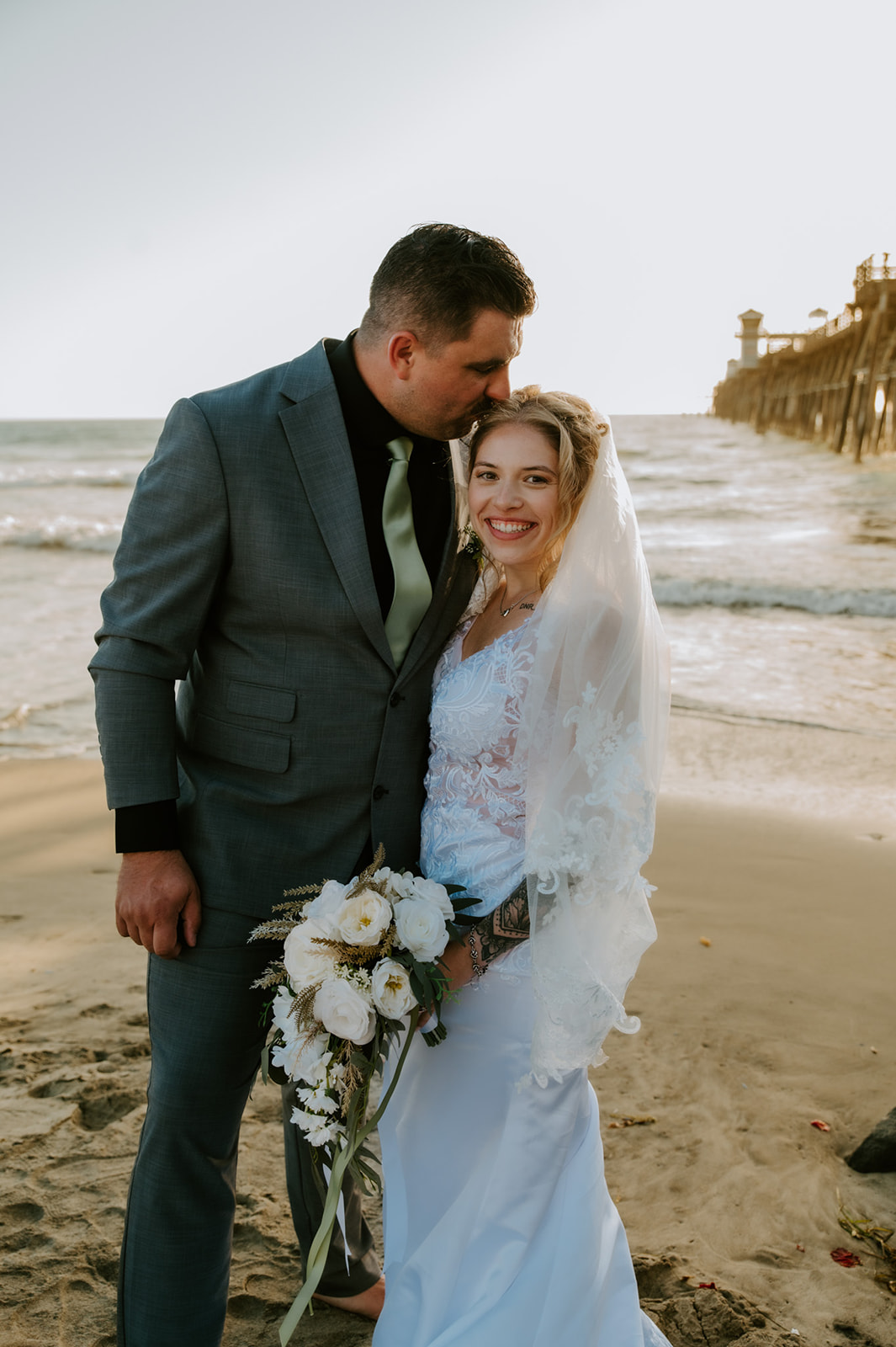 Bride smiling as groom kisses her forehead on the beach, a heartfelt moment from their California elopement