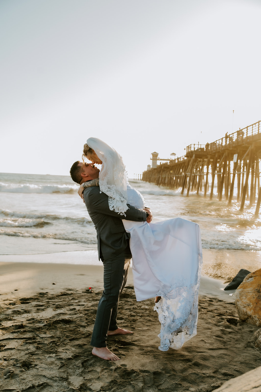 Groom lifting the bride in the sand near an Oceanside pier during sunset, capturing carefree beach elopement photos after a family-inclusive California elopement.