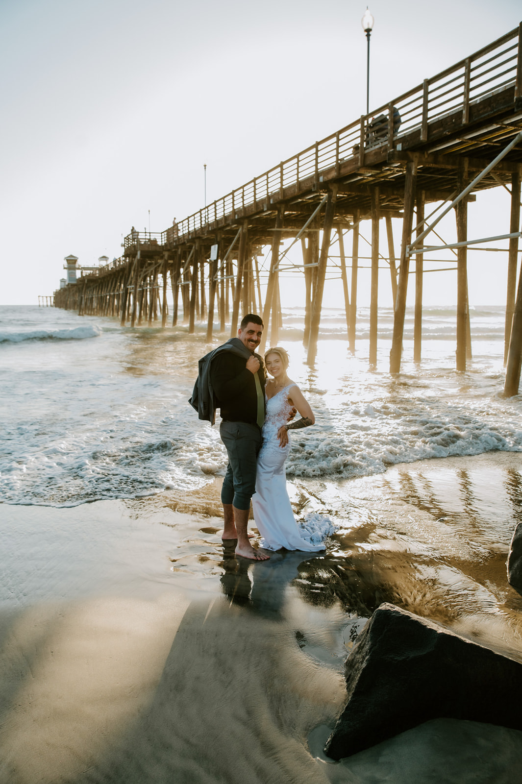 Bride and groom standing together in the surf near an Oceanside pier, ending the day with relaxed, candid beach elopement photos.