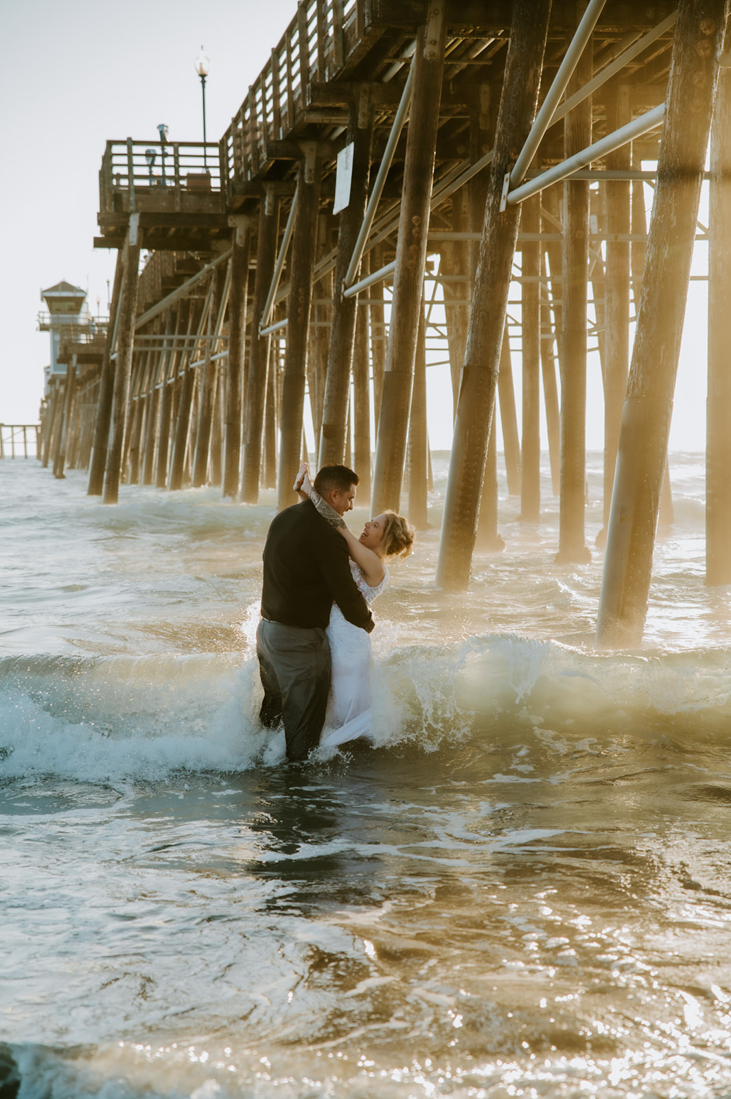 Bride and groom embracing in the ocean under the pier after their family ceremony, showing how to plan an elopement with family while leaving space for private moments.