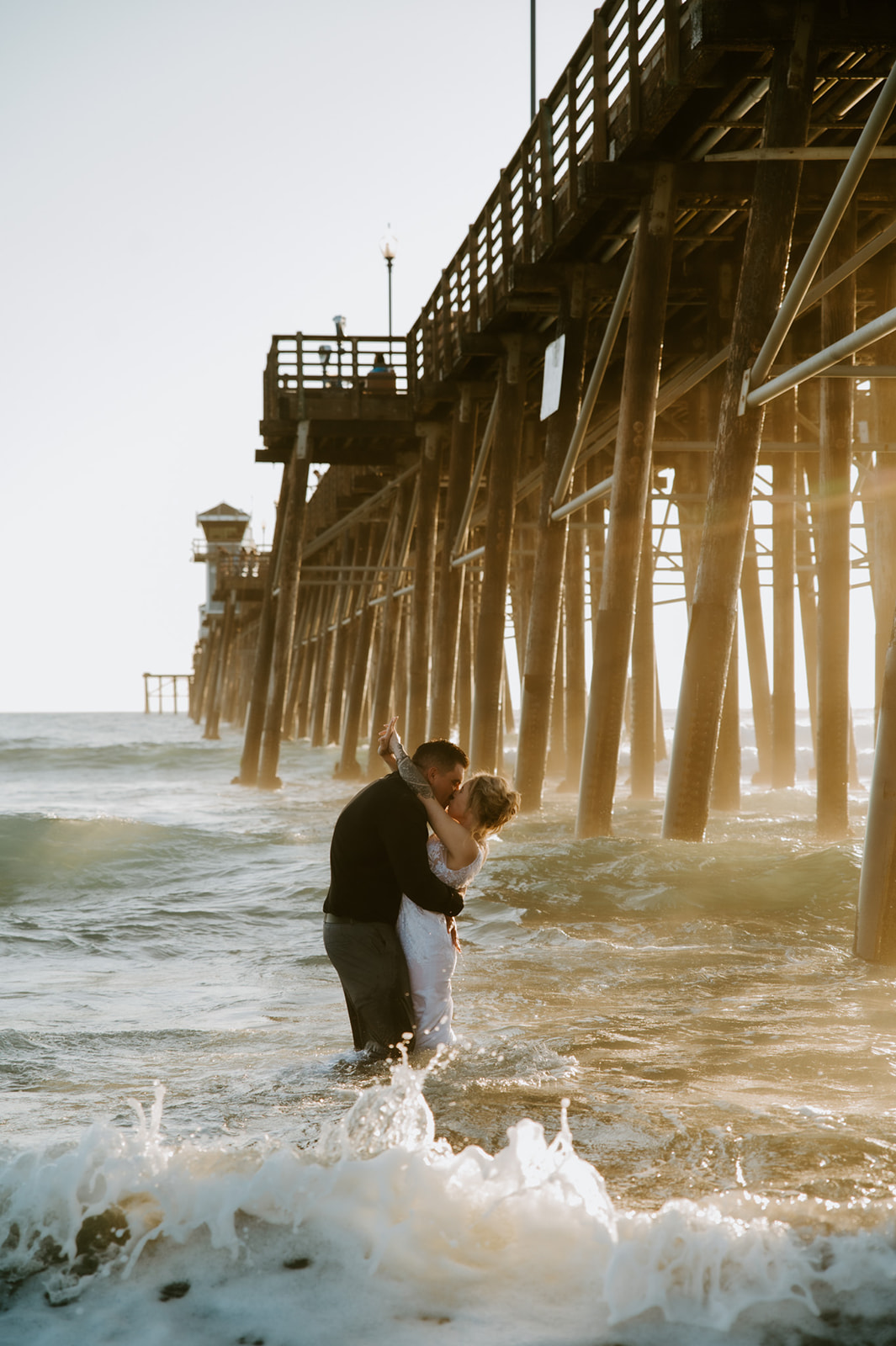 Couple embracing in the waves beneath the pier, showing how to plan an elopement with family and still lean into adventure.