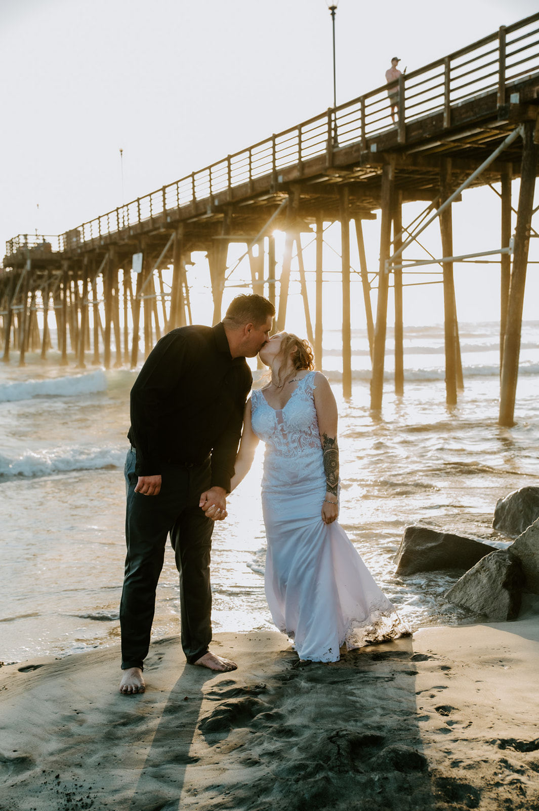 Couple kissing on the shoreline with the pier behind them during sunset beach portraits.
