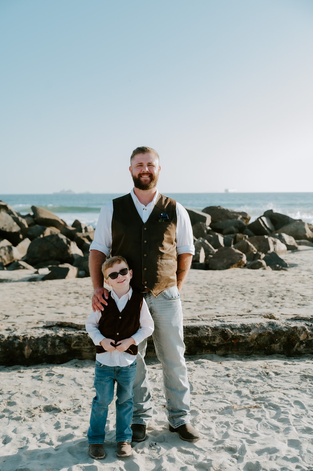 Groom wearing brown vest and jeans standing with young son wearing sunglasses and smiling.