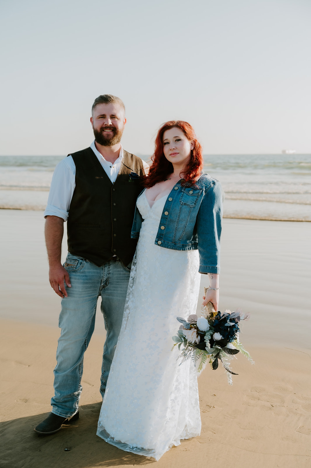 Bride and groom standing together on the shoreline after their beach elopement, holding a bouquet and smiling toward the camera, photographed by a california elopement photographer in soft coastal light.