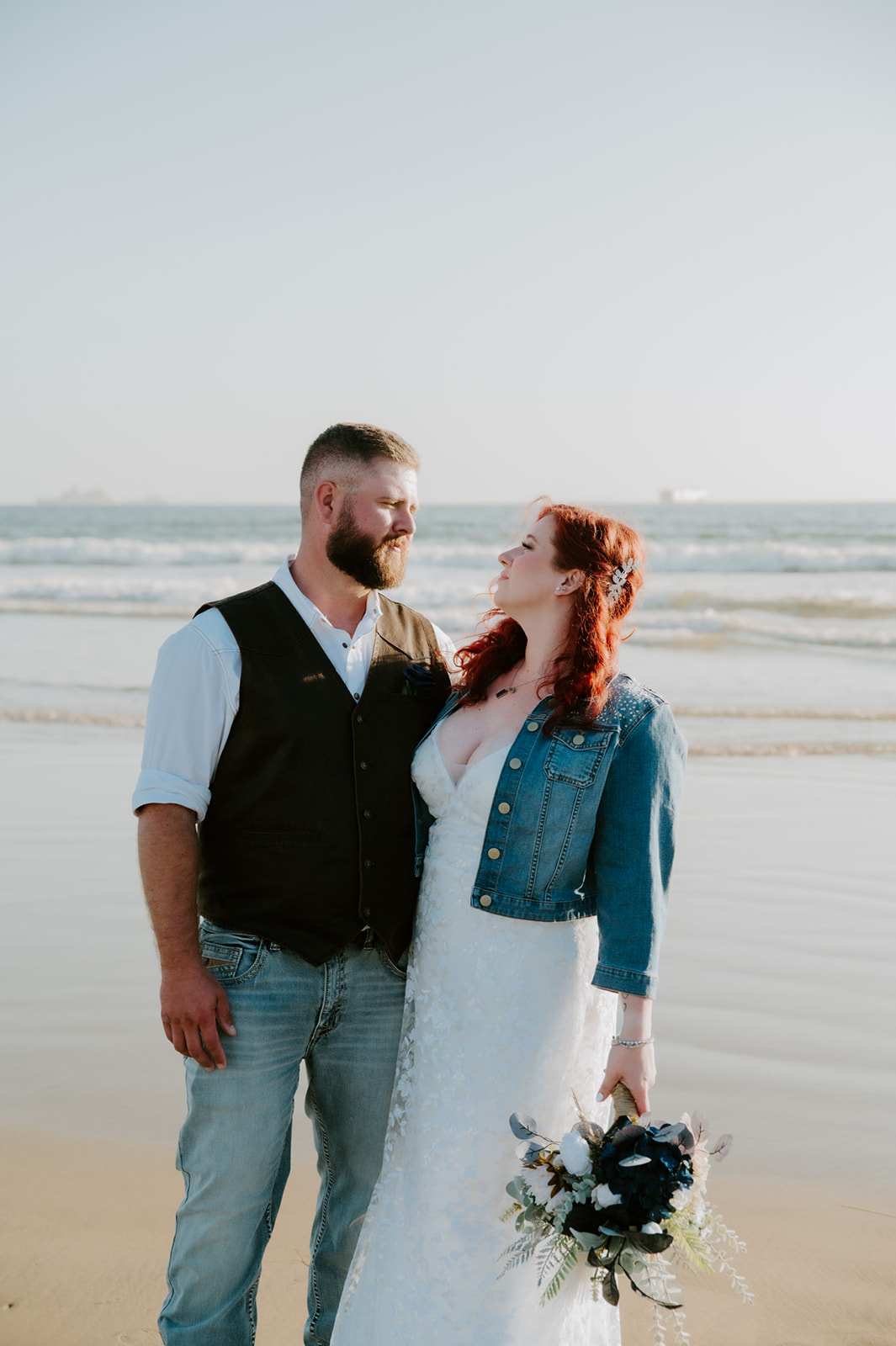 Bride and groom facing each other on the beach during their California elopement ceremony, city skyline faintly visible behind them.