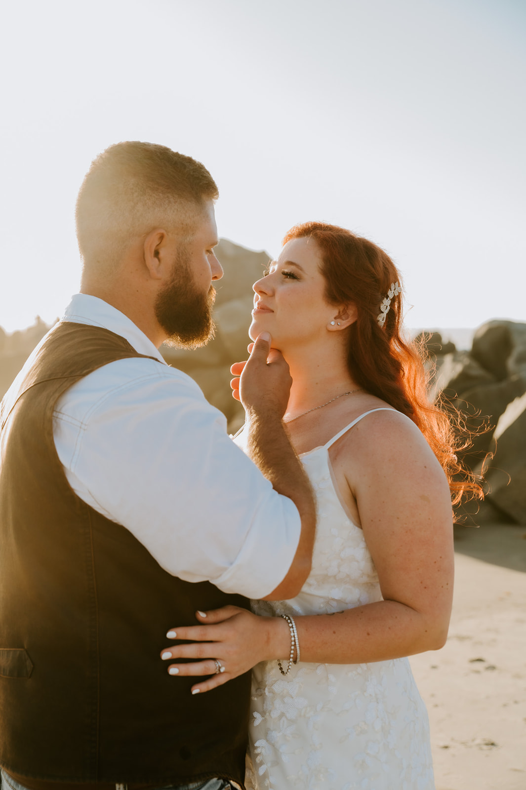 Groom gently touching the bride’s face during golden hour beach portraits with ocean light wrapping around them