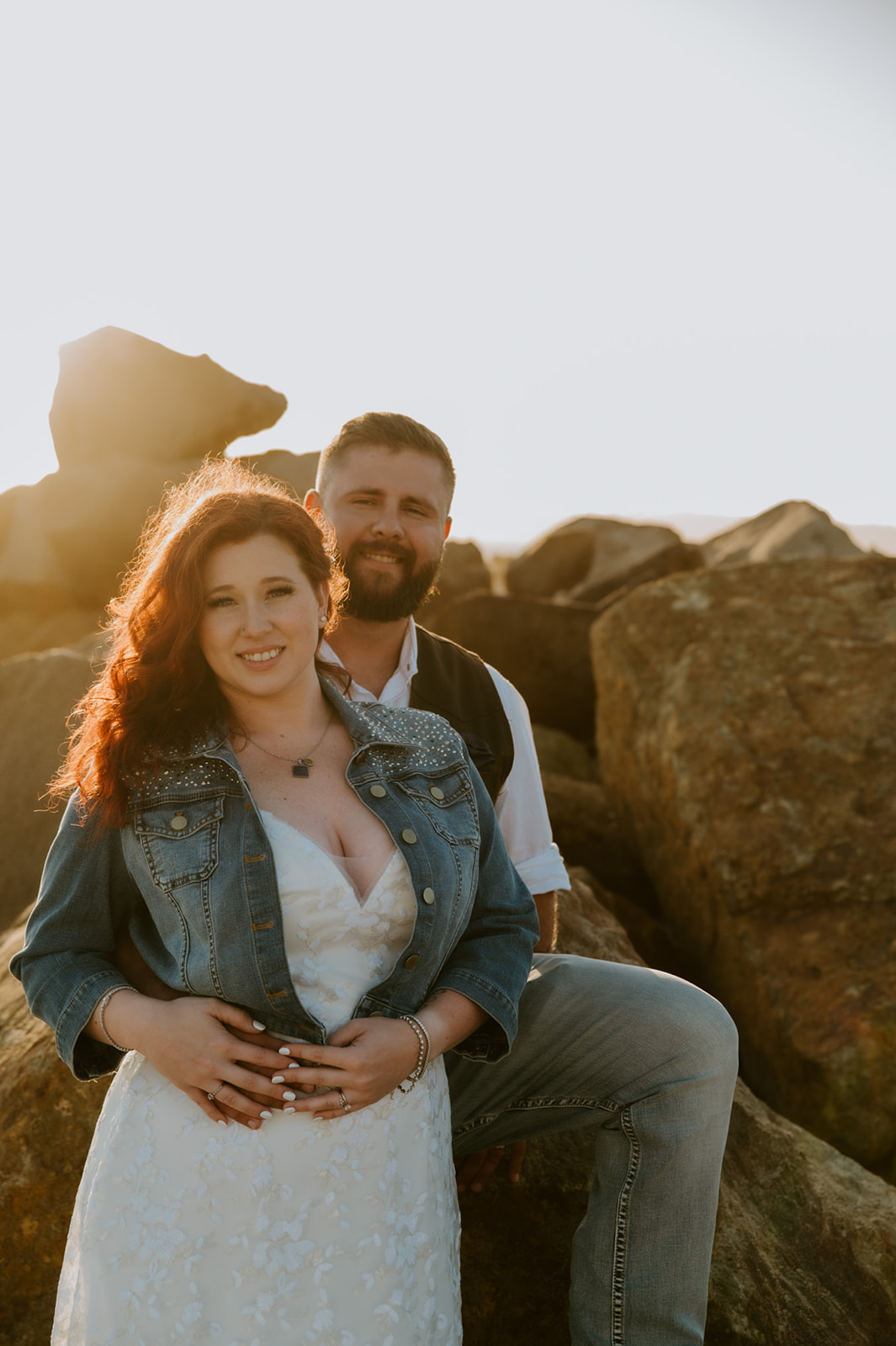 Couple sitting together on large coastal rocks at sunset, smiling softly during an intimate beach elopement