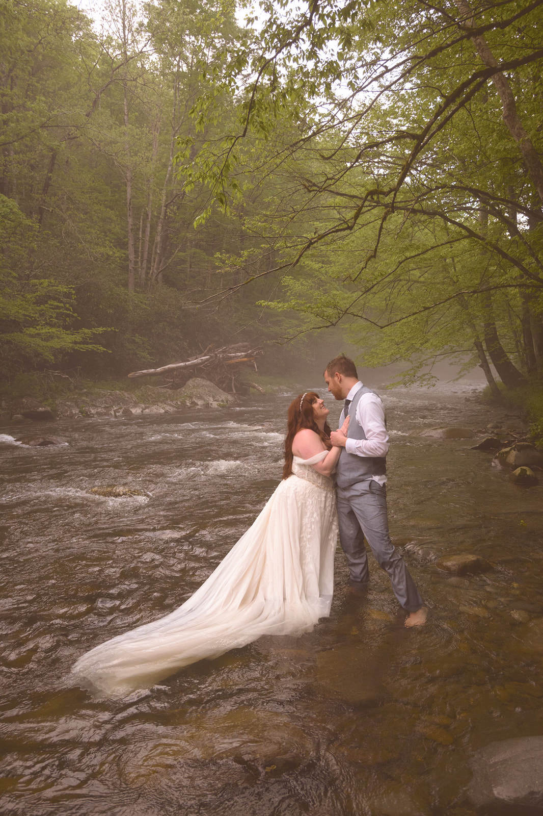Bride and groom standing barefoot in a foggy creek, holding each other as the river flows around them during an adventurous elopement session.