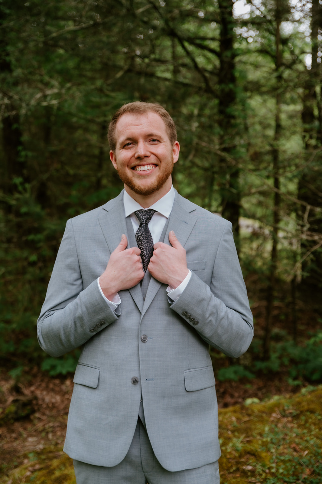Groom smiling in a light gray suit while standing in a forest setting before the ceremony.