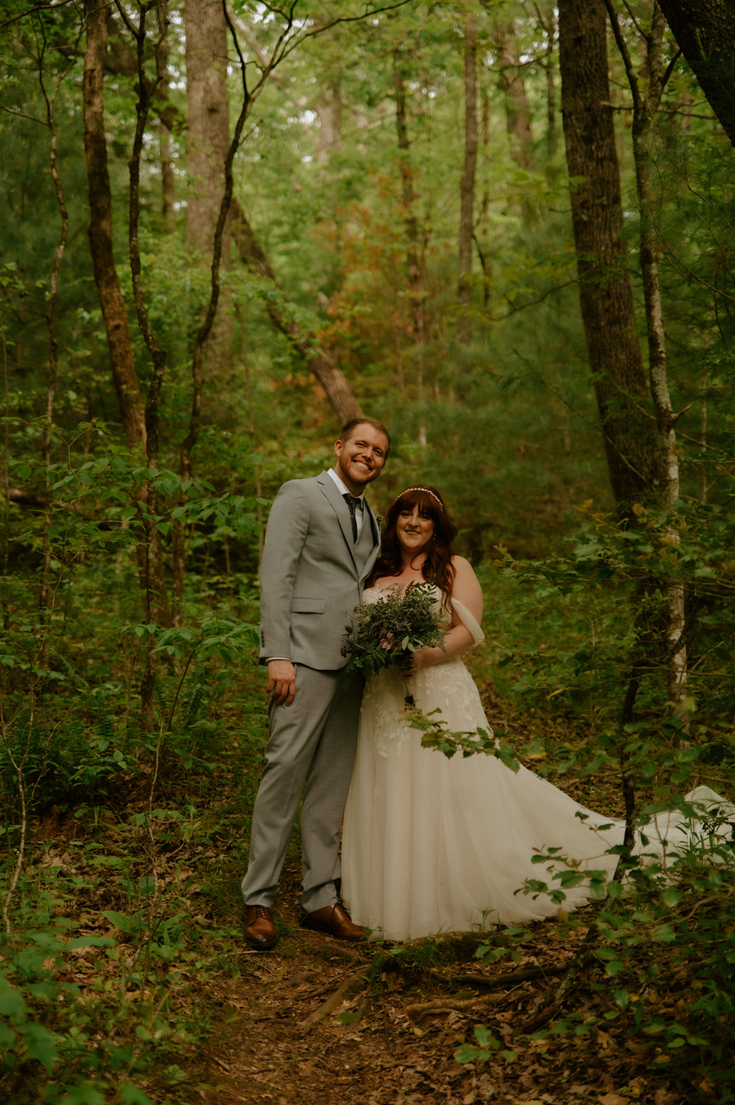 Bride and groom standing together in a wooded forest clearing, surrounded by trees and soft natural light.