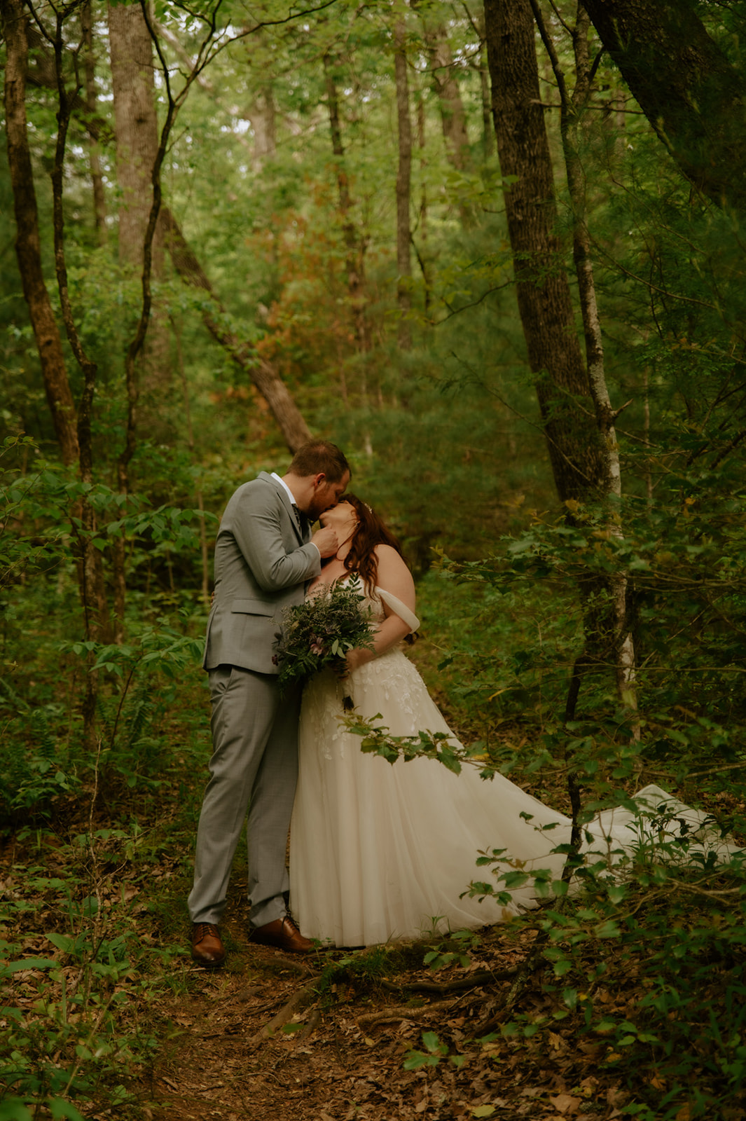 Bride and groom kissing in a wooded area with soft natural light filtering through the trees