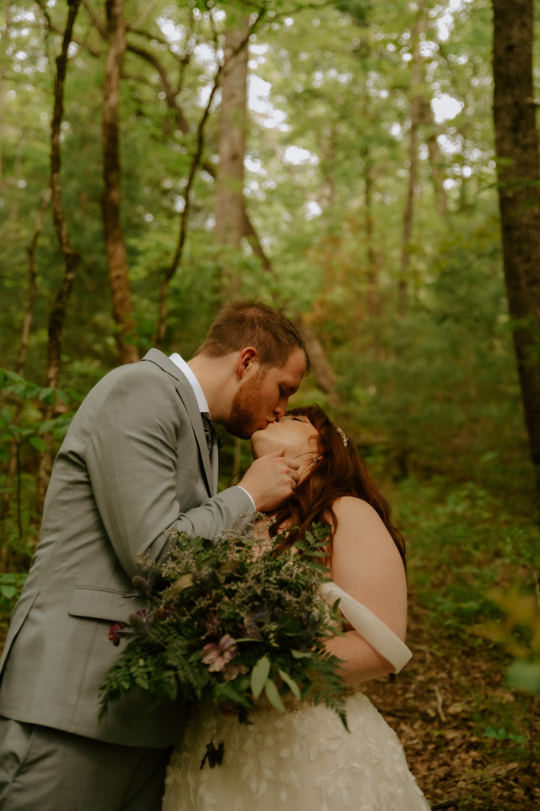 Couple sharing a kiss beneath the trees while holding a bouquet during their elopement session.