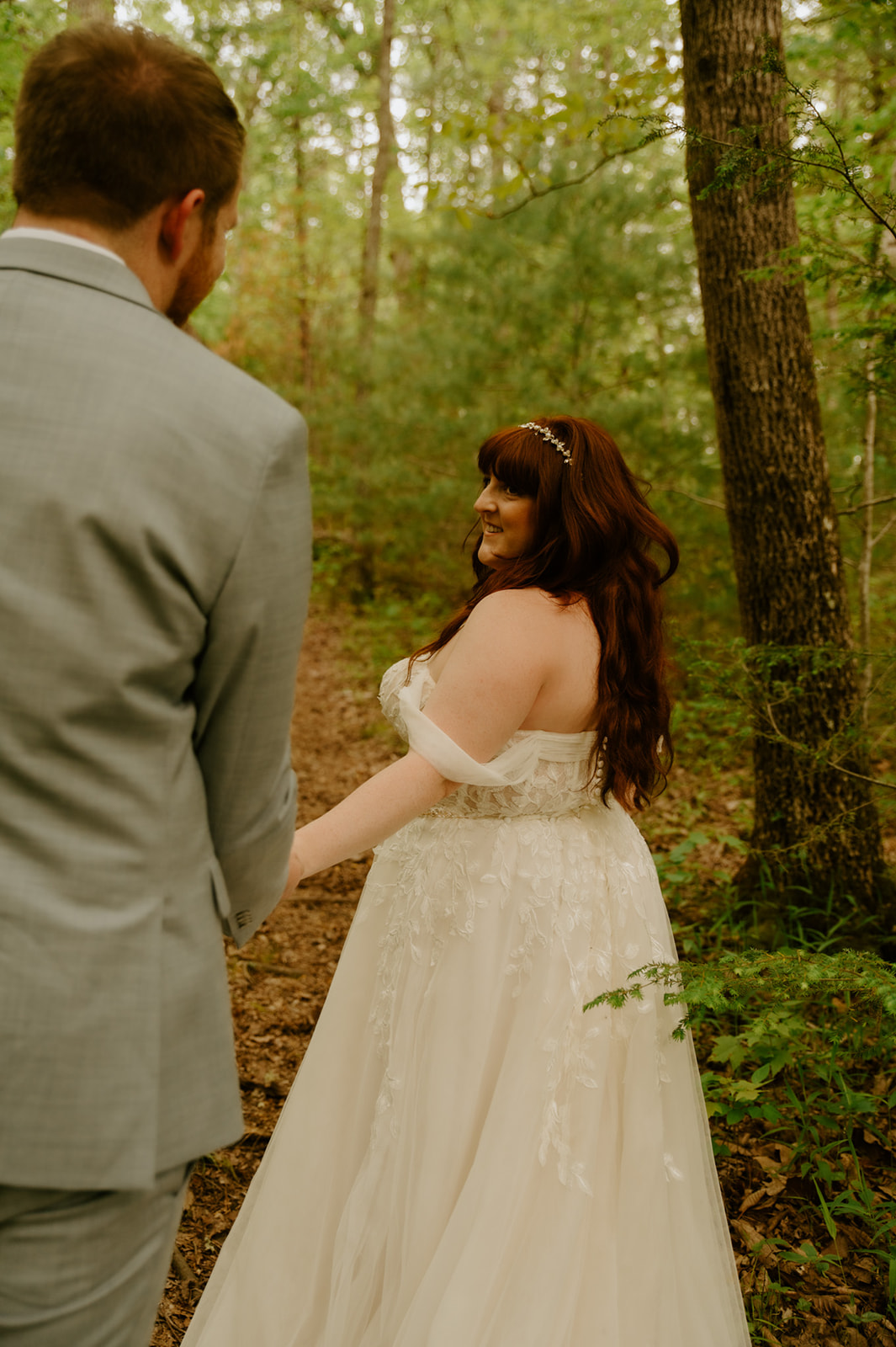 Groom reaching for the bride’s hand as she looks back smiling during forest portraits.