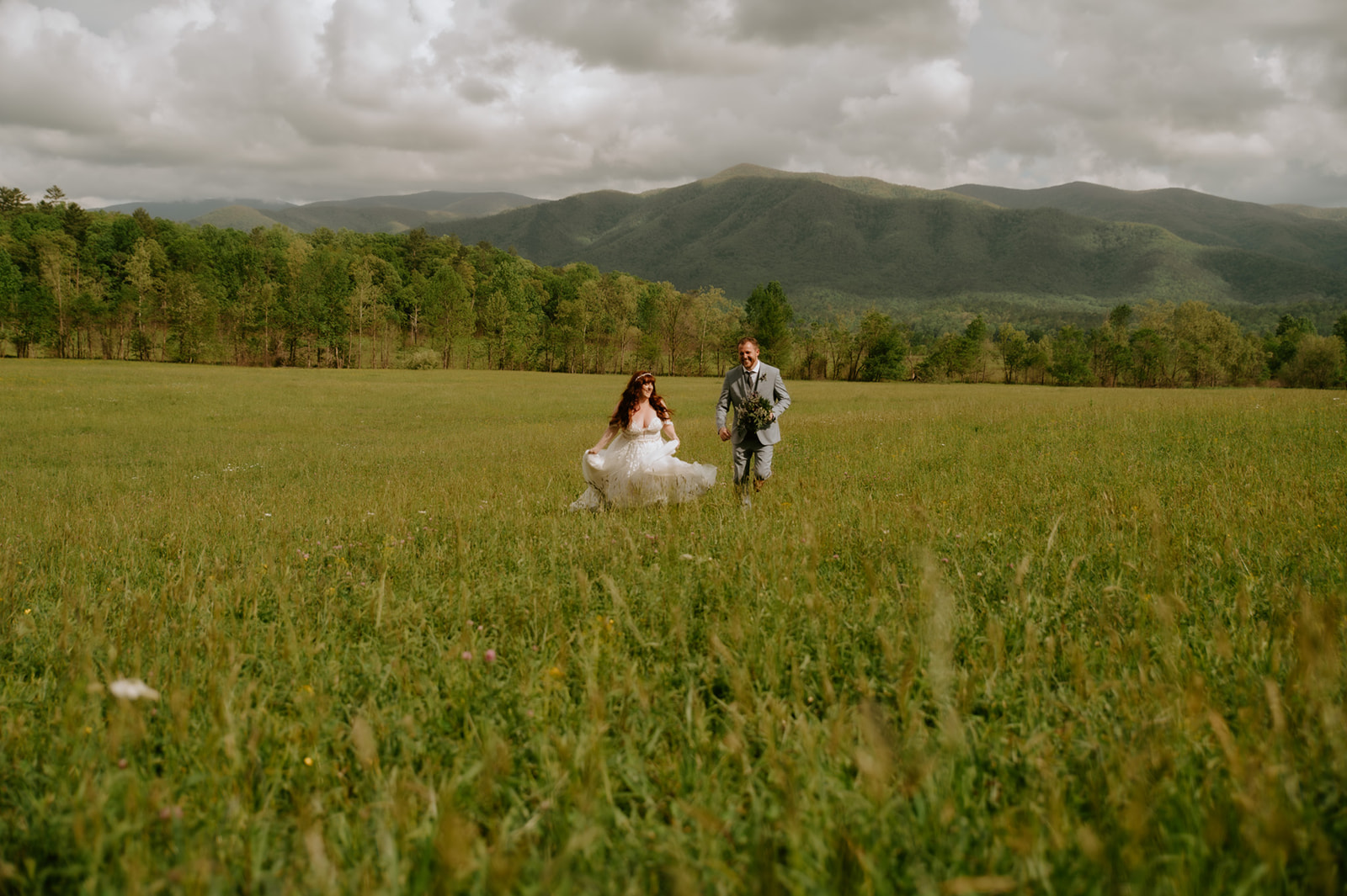 Wide meadow portrait of the bride and groom running together with the Smoky Mountains stretching behind them.