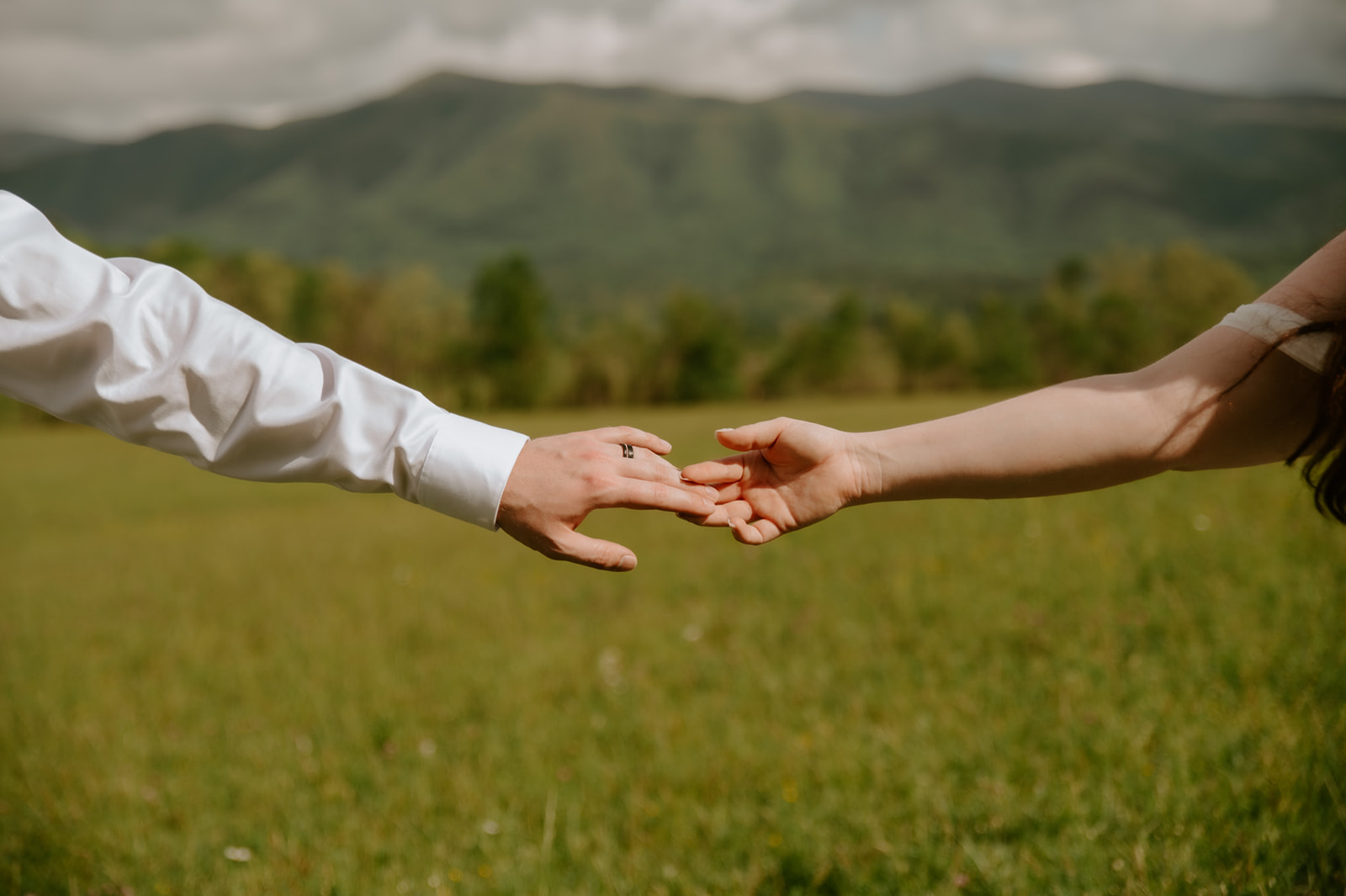 Close-up of bride and groom reaching for each other’s hands in a sunlit mountain meadow