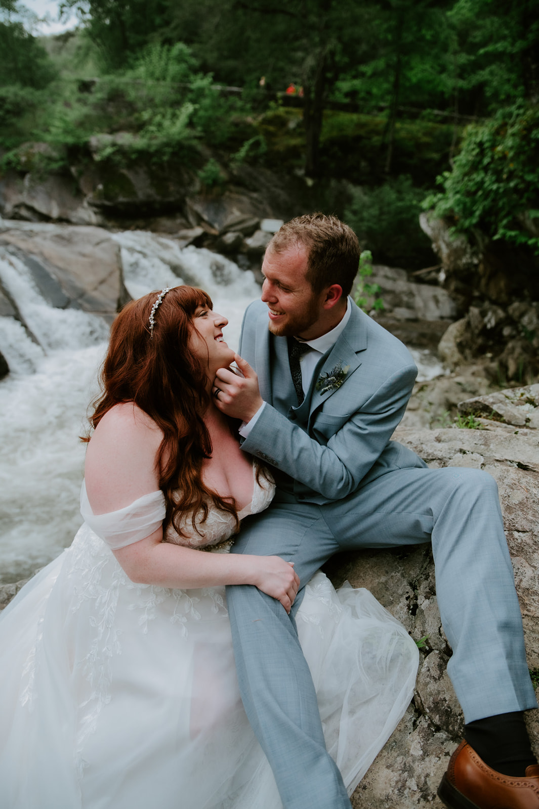 Bride and groom sitting on rocks near a waterfall, laughing together during their adventure elopement