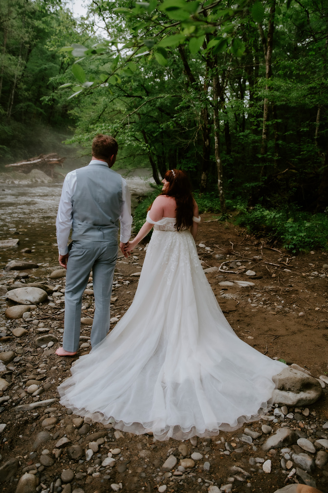 Bride and groom walking hand in hand along a rocky riverbank, dress trailing behind her.