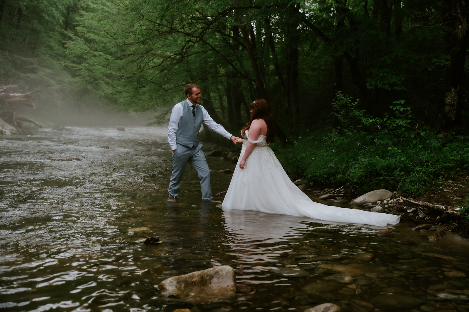 Bride and groom standing in the river together, holding hands as fog drifts through the forest during a Smoky Mountain elopement.