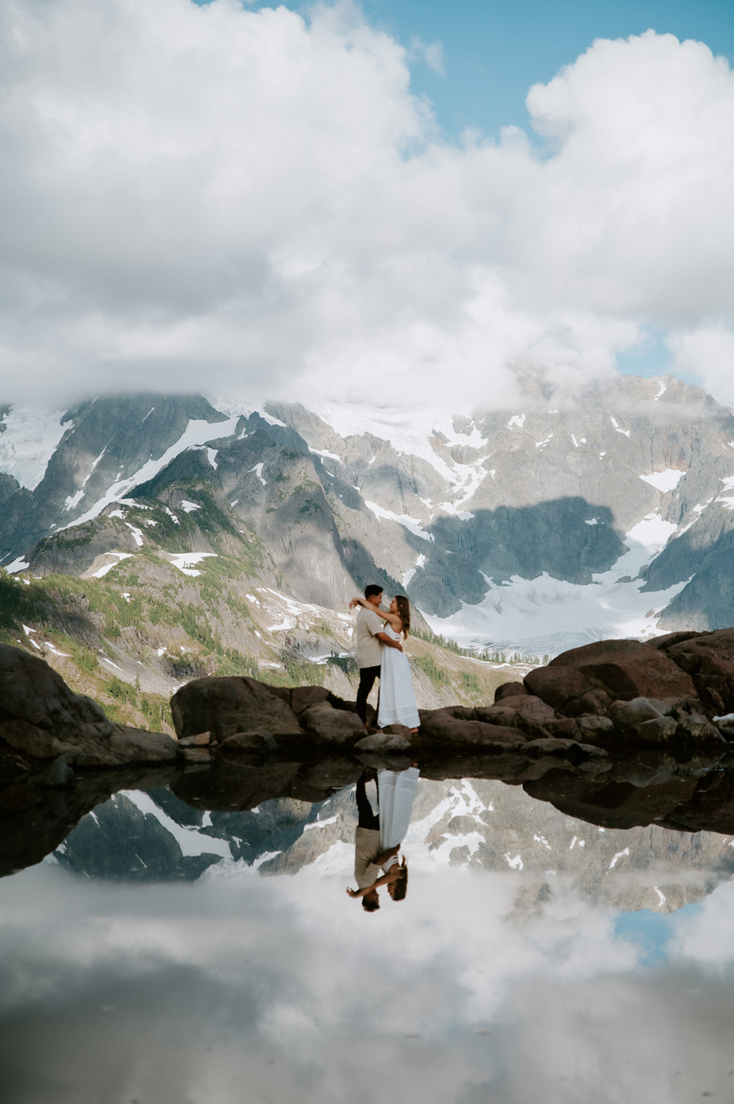 Wide landscape photo of a couple standing together on rocks by an alpine lake while they elope in Washington, mirrored by surrounding peaks.