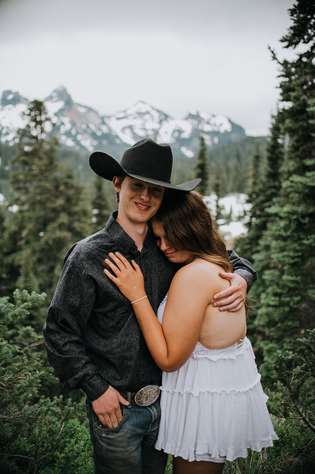 Couple cuddling among evergreen trees with snow-capped mountains behind them while they elope in Washington in the Pacific Northwest.