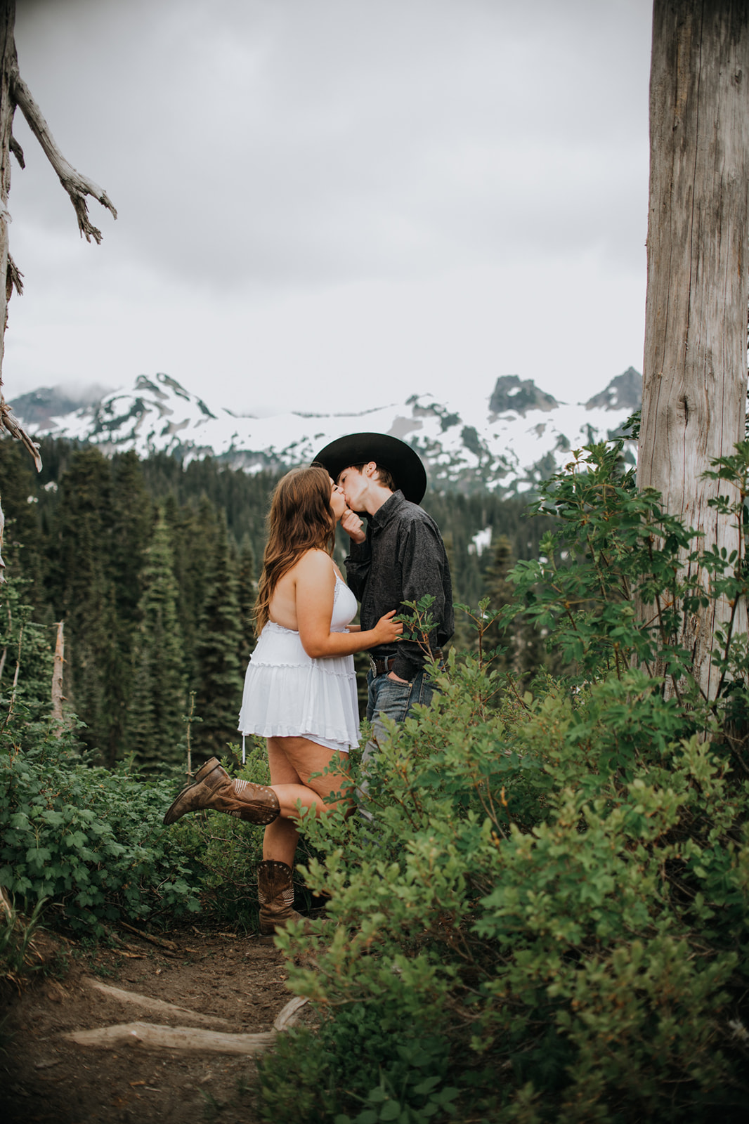 Couple kissing in a lush alpine forest clearing with snow-capped peaks visible through the trees during an intimate elopement session.