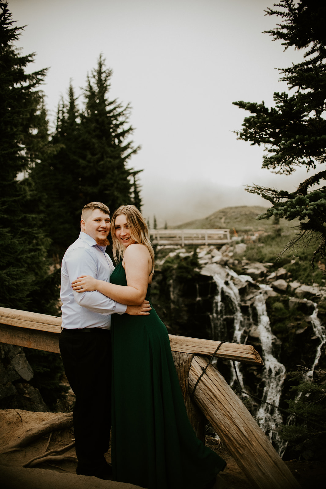 Couple standing on a wooden overlook near a waterfall in formal attire while they elope in Washington on a misty mountain day.