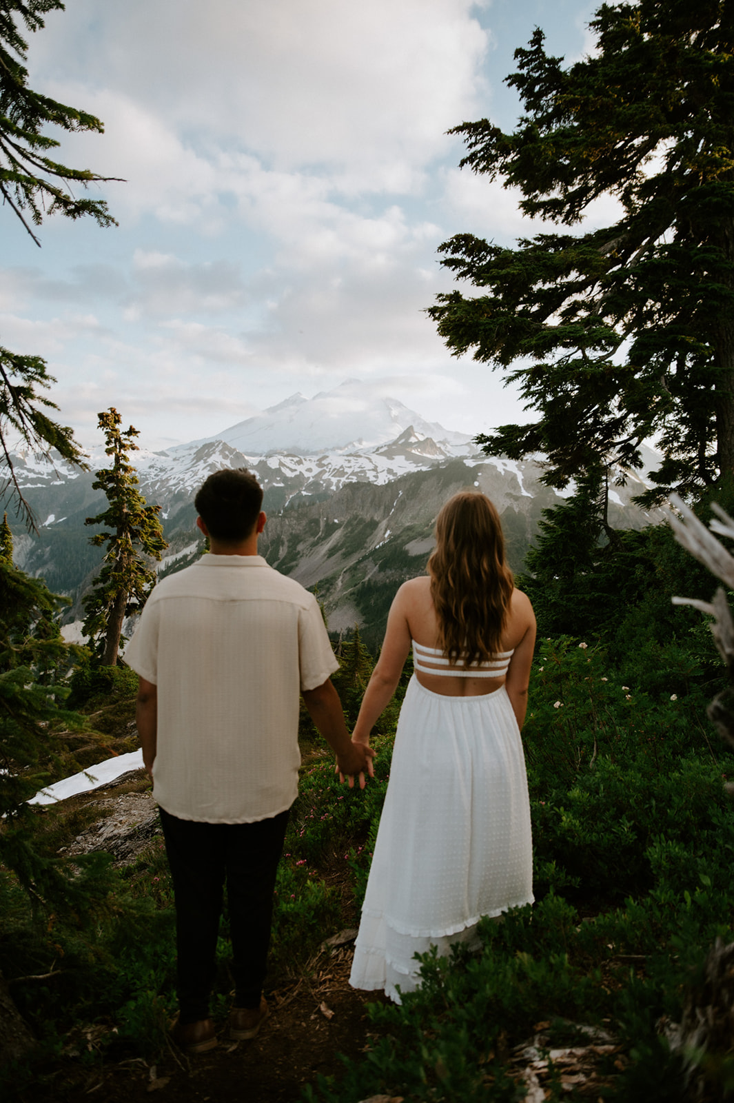 Couple holding hands on a forested mountain trail with snowy North Cascades peaks in the distance