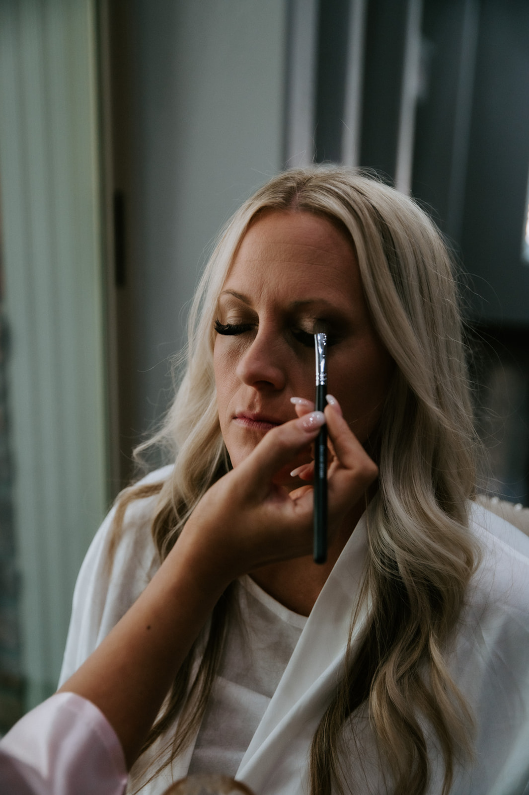 Bride having her makeup done by natural window light during a slow cabin morning before her elopement.