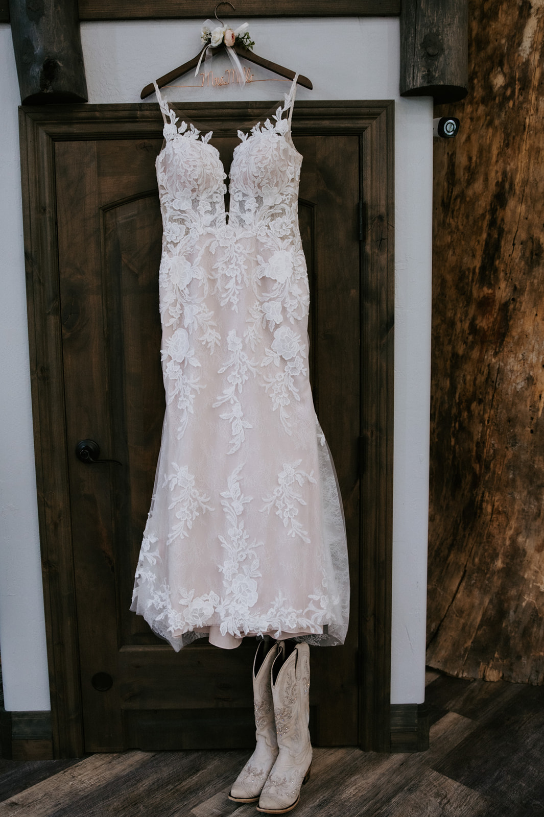 Lace wedding dress hanging on a rustic wooden door with cowboy boots underneath, styled for an Arizona forest wedding.