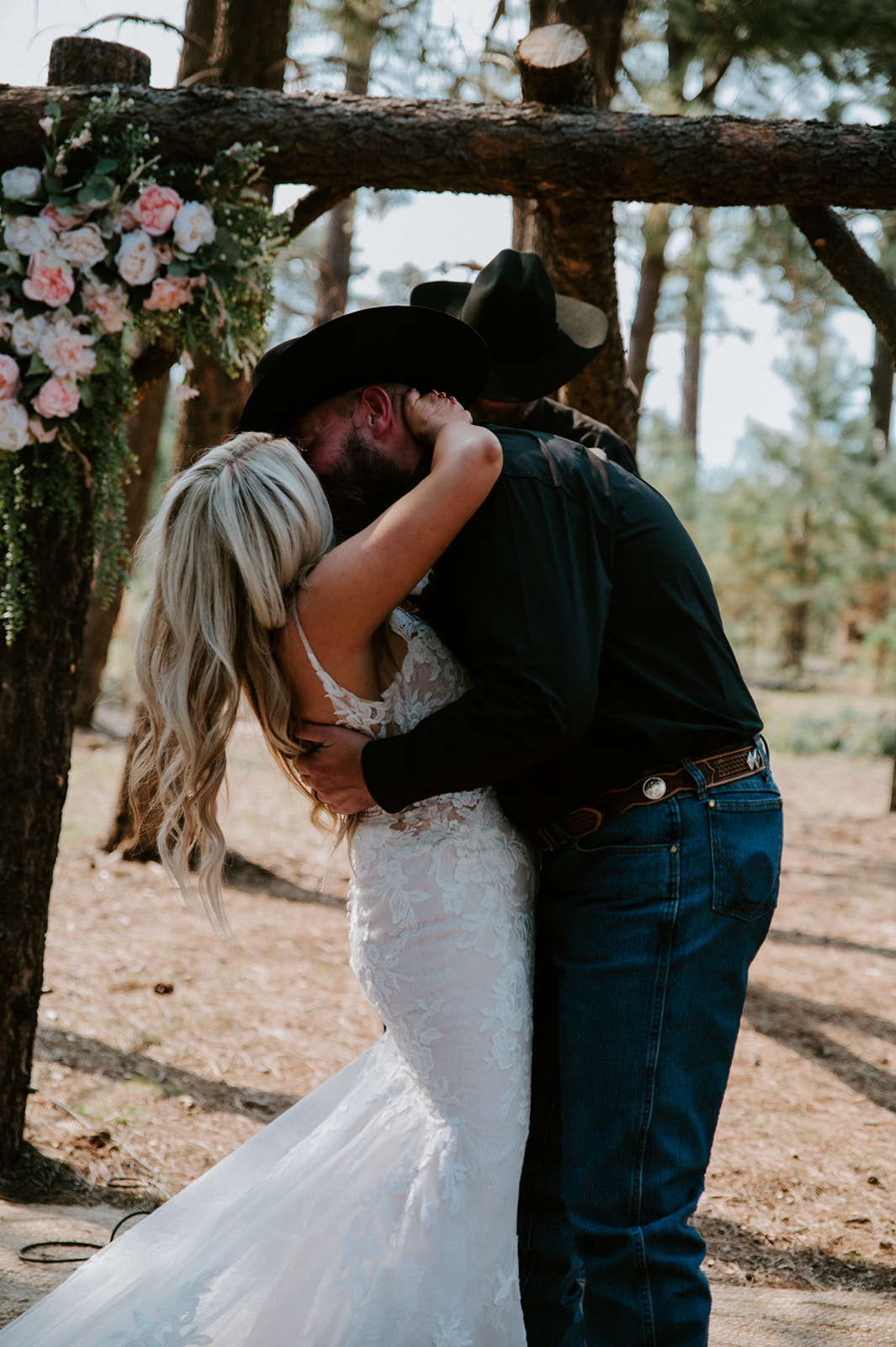 Bride and groom sharing their first kiss under a floral-covered wooden arch in the Arizona forest.