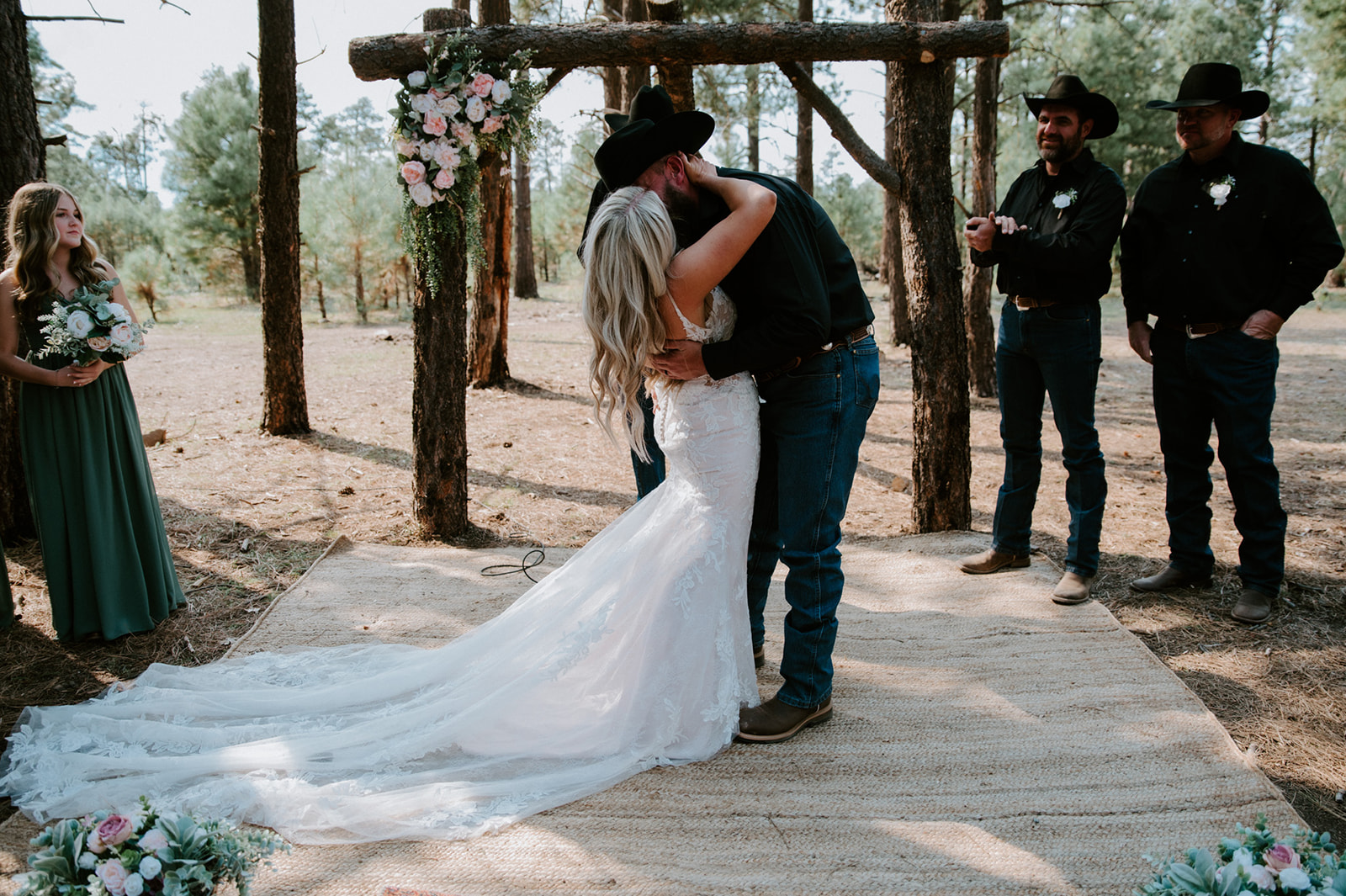 Bride and groom sharing their first kiss under a floral-covered wooden arch in the Arizona forest.