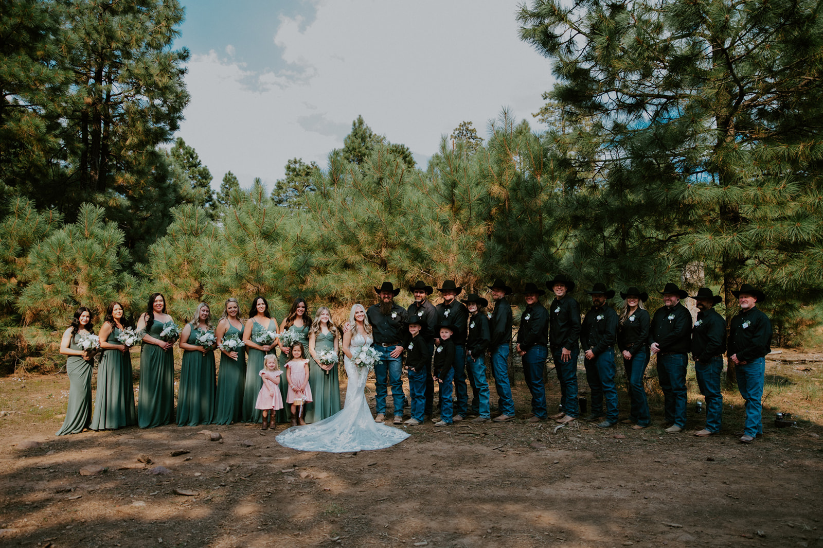 Large wedding party portrait in the pines with bridesmaids in green dresses and groomsmen in black western attire during an Arizona forest wedding.
