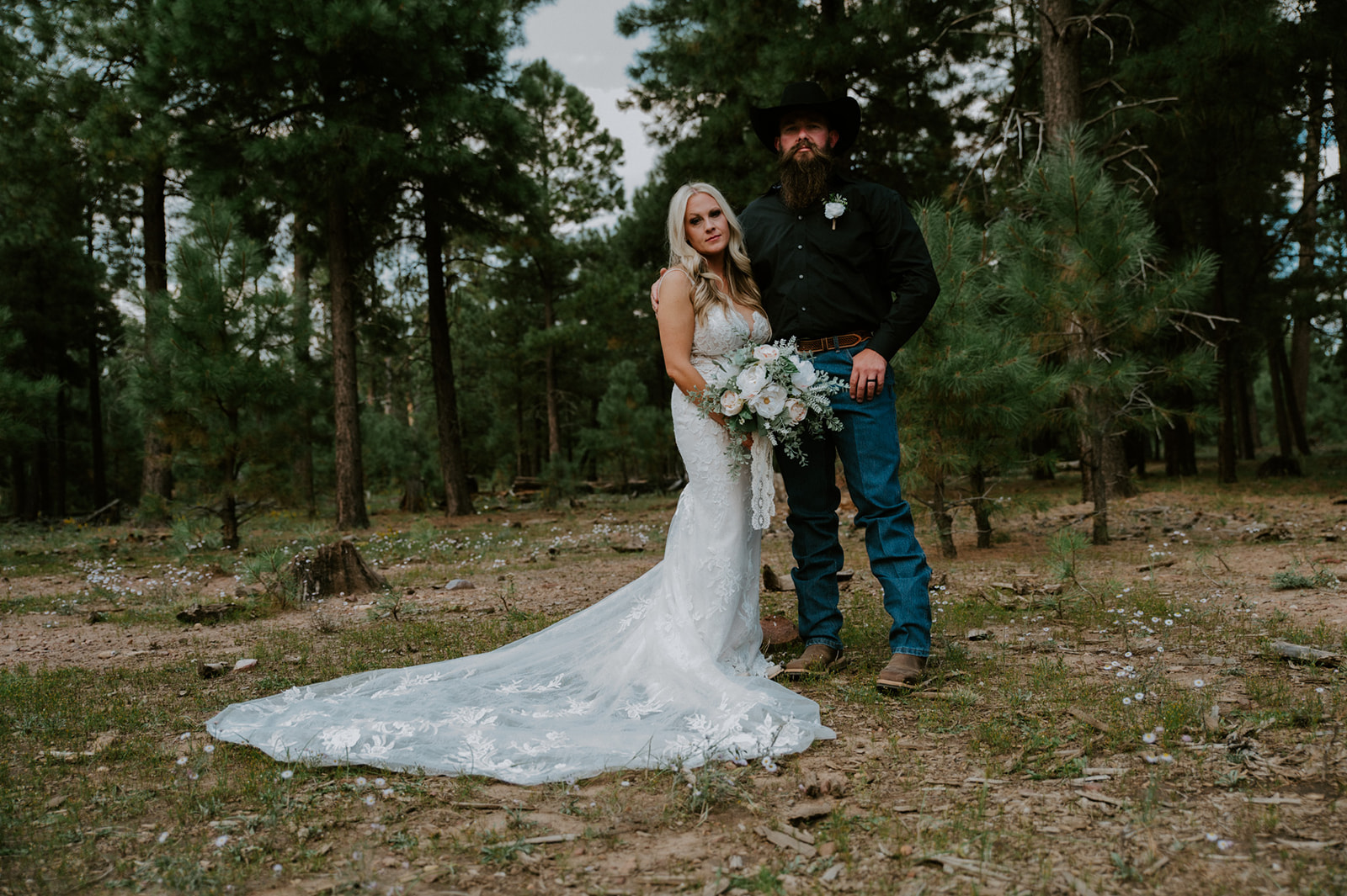 Bride and groom posing together in a pine forest clearing with her lace train spread across the ground during their Arizona elopement.