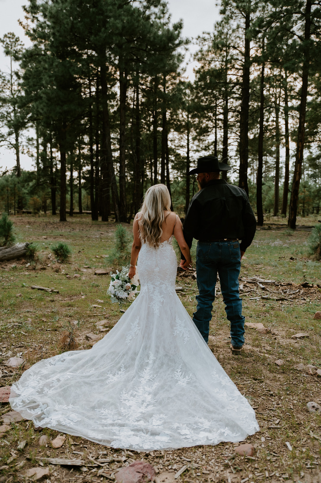 Bride and groom walking hand in hand through tall pine trees with her lace train trailing behind during their elopement in Arizona.