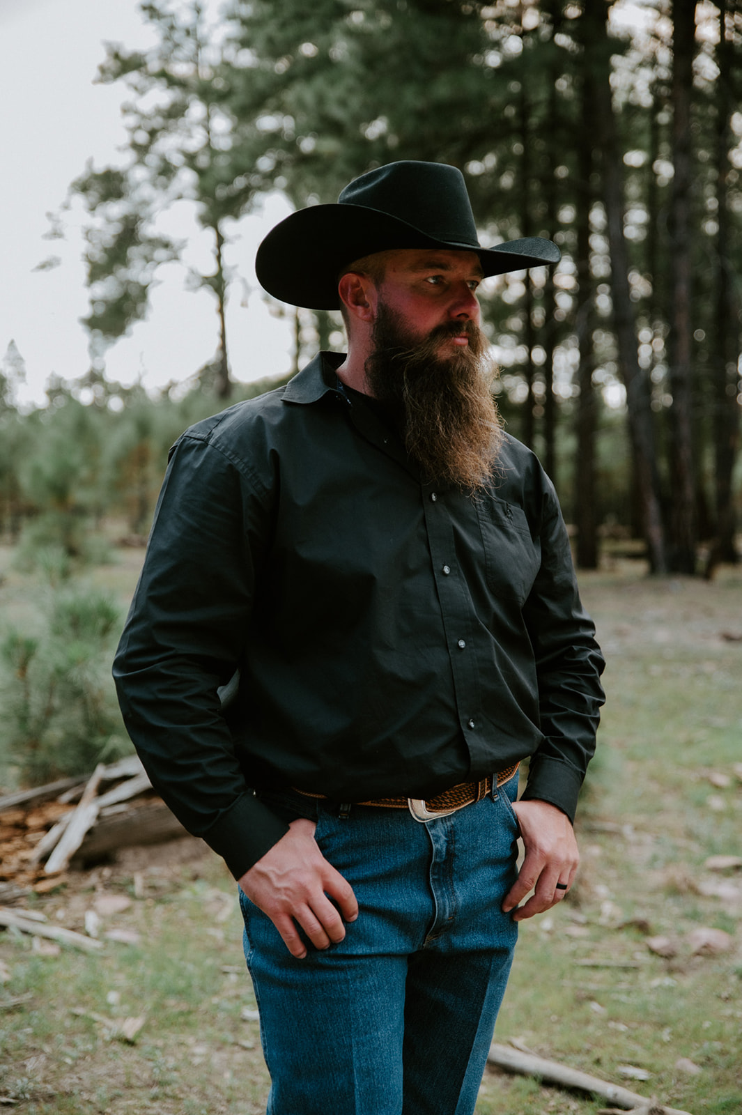 Groom in black cowboy hat and western attire standing in a pine forest before his ceremony to elope in Arizona.