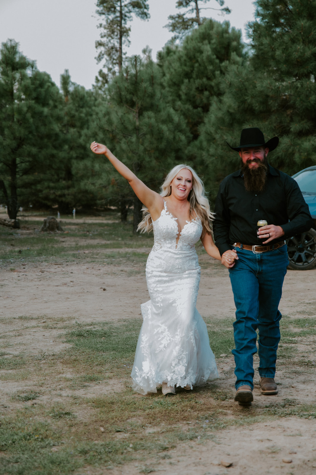 Newlyweds walking hand in hand through a forest clearing with bride cheering and groom holding a drink after they elope in Arizona.
