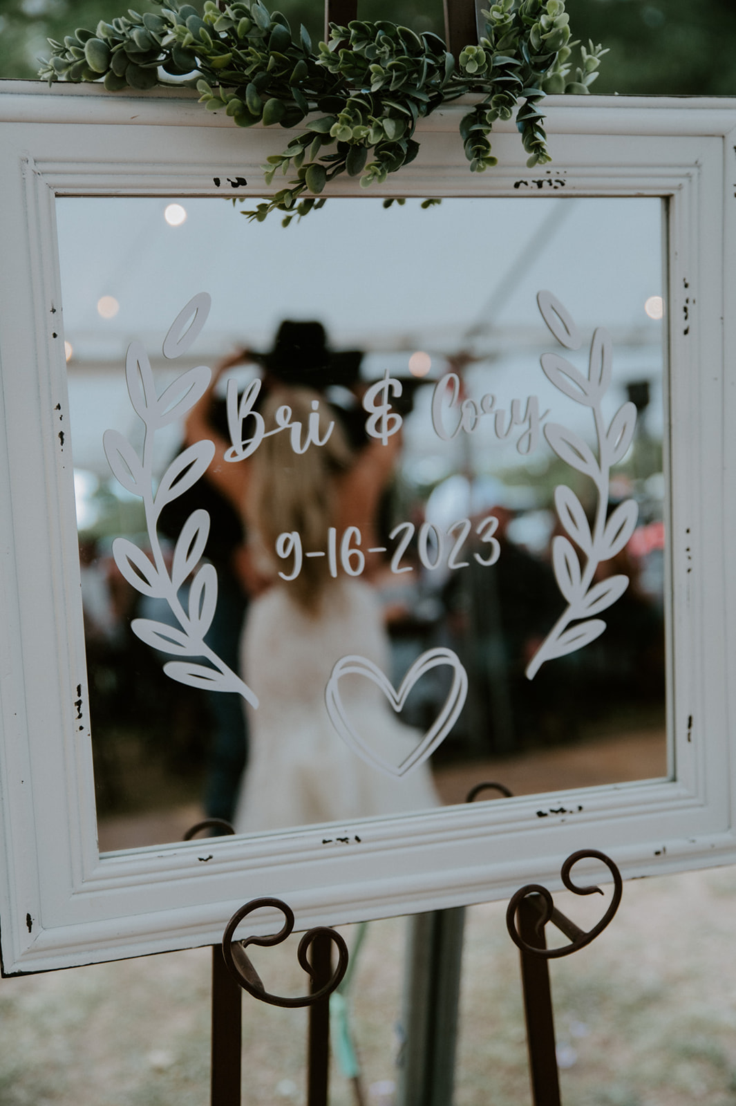 Wedding welcome mirror sign reading “Bri & Cory 9-16-2023” with greenery above and the couple blurred in the reflection at their forest reception in Arizona.