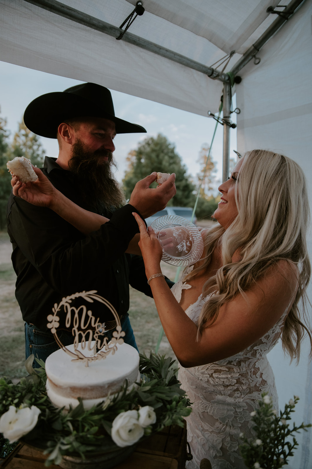 Bride and groom laughing while feeding each other cake under a reception tent after they elope in Arizona.