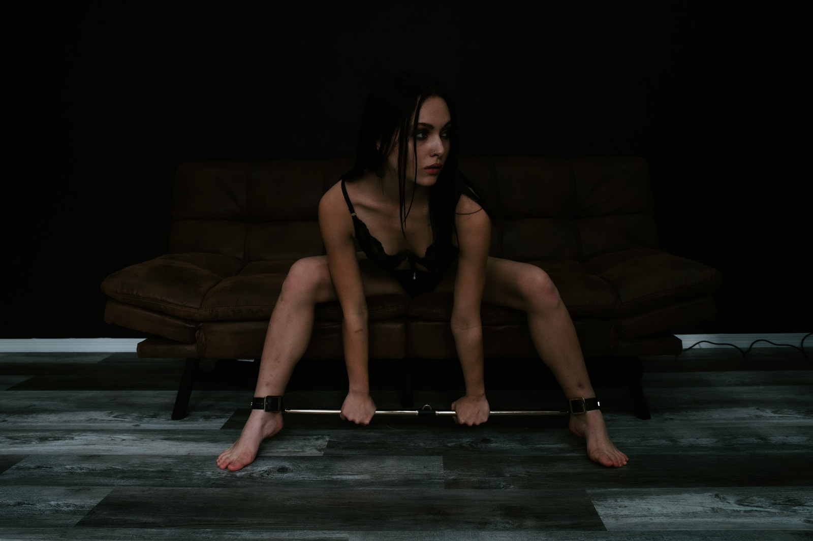 Bride seated low on a couch gripping a bar during a boudoir photo shoot, strong posture and direct gaze in moody light