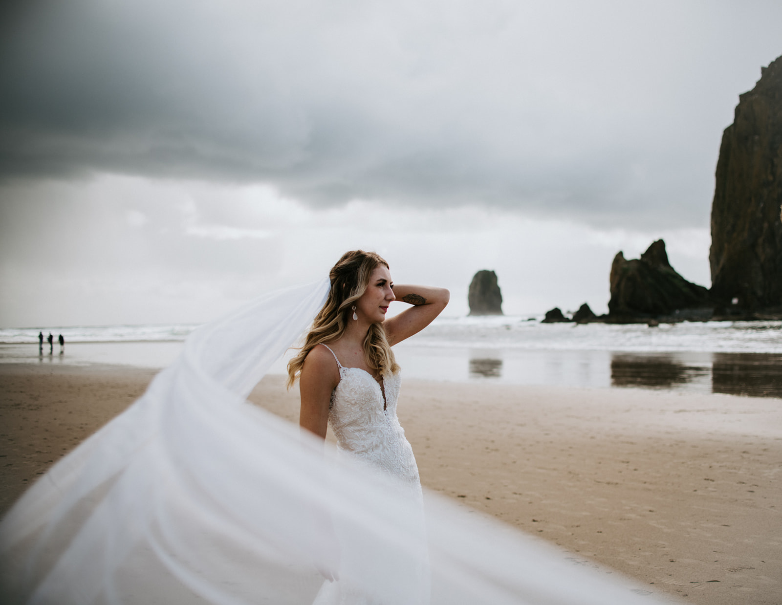 Bride touches her veil to secure it from the wind blowing it away while she looks off into the ocean. 